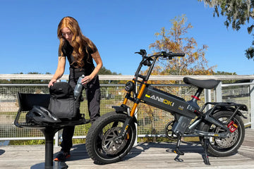 A grey and gold Aniioki A8 fat tire e-bike parked on an outdoor wooden boardwalk under a clear blue sky, with a Caucasian woman packing a black backpack next to it
