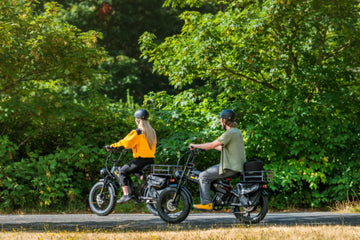 A Caucasian man and woman riding black Aniioki AQ177 48V long range electric bikes on a paved park trail surrounded by green trees